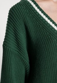 Pull vert en tricot avec un col en V, présentant un côtes texturé et un accent rayé blanc le long du col. Tissu doux et épais.