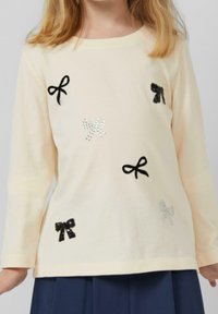 Jasnobeżowa koszulka z długim rękawem z czarnymi i srebrnymi aplikacjami w kształcie kokardek oraz akcentami z kryształków. Miękka bawełniana tkanina o luźnym kroju.