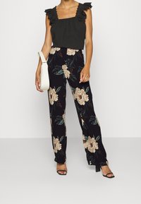 Czarny top bez rękawów z falbaniastymi koronkowymi ramiączkami, w połączeniu z czarnymi spodniami o szerokich nogawkach w kwiatowy wzór, z beżowymi i zielonymi kwiatami. Biała torebka i szpilki.