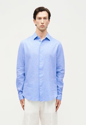 Muž oblečený v svetlomodrej košeli s dlhým rukávom a bielych nohaviciach stojaci pred jednoduchým svetlým pozadím.