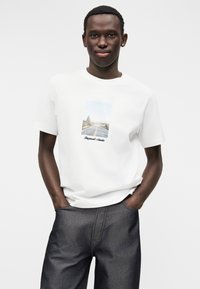 Λευκό βαμβακερό t-shirt με εκτύπωση γραφικών τοπίου και κείμενο από κάτω. Το μοντέλο το συνδυάζει με σκούρο τζιν παντελόνι, με τα χέρια στις τσέπες.