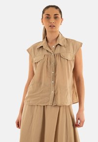 Camisa sin mangas beige, con botones y cuello, con bolsillos frontales y detalle de fruncido. Fabricada en una tela ligera y texturada.