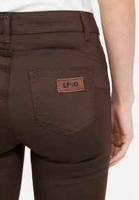 Jeans stretch marroni con un design aderente, tasca posteriore con etichetta cucita e texture liscia. Leggero lucido visibile sul tessuto.