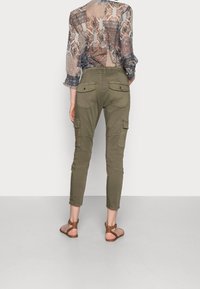 Olive green cargo pants with tapered legs, cargo pockets on thighs, and back pockets. Paired with a sheer patterned blouse and brown sandals.