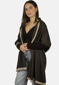 Sciarpa marrone con rifiniture metalliche dorate, design ampio e comodo, materiale strutturato, indossata sopra una maglia scura e abbinata a jeans chiari.