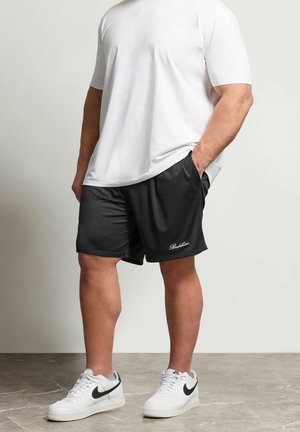 Mand iført hvid kortærmet skjorte, sorte sportsshorts med sidelommer og hvide Nike sneakers, stående på et flisebelagt gulv.
