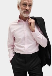 Světle růžová košile na knoflíky s dlouhými rukávy je spárována s tmavě šedými kalhotami. Na jednom rameni je přehozený černý blazer.