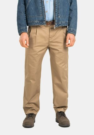 Man standing wearing beige pleated trousers, brown belt, blue denim jacket, light blue shirt, and dark brown shoes against white background.