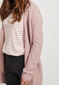 Světle růžový žebrovaný cardigan s dlouhými rukávy, nošený přes bílou a světle růžovou pruhovanou košili, v kombinaci s černými kalhotami.