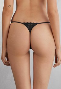 Zwarte kantenslips met golvende randen, minimale bedekking en dunne bandjes. Gladde textuur tegen de huid, die een delicate uitstraling benadrukt.