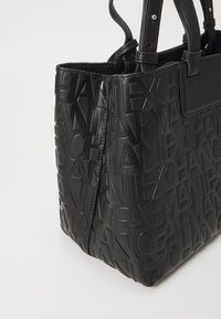 Svart tote bag med preget logo, strukturert form, glatt tekstur og doble topphåndtak for bæring.