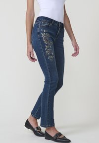 Denim kavbojke temno modre barve z vezenimi cvetličnimi in paisley motivi v zlati in rdeči barvi, s pasom srednje višine in ravnim krojem.