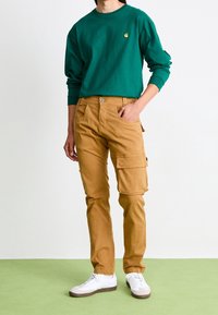 Grön långärmad skjorta, bruna cargobyxor med flera fickor, vita sneakers. Slätt tyg, avslappnad passform och neutral färgpalett.