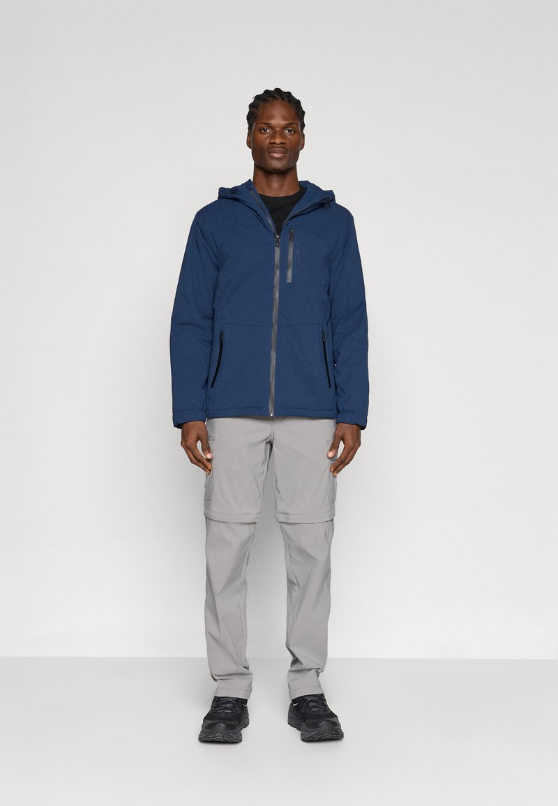 Chaqueta con cremallera color azul marino y capucha, con un bolsillo en el pecho y bolsillos laterales, combinada con pantalones gris claro y zapatos negros.