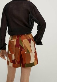 Multicolored patterned shorts with geometric shapes in warm tones, featuring large pockets and a folded hem, paired with a sheer black top.