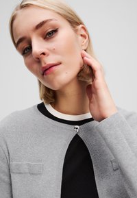 Cardigan grigio a maglia con scollo tondo e chiusura a bottoni, caratterizzato da un accento bianco e nero sulla parte interna del colletto e una spilla decorativa.
