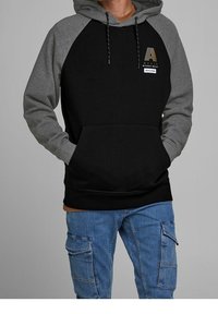 Uomo che indossa una felpa nera e grigia con tasche frontali e jeans cargo blu, mani nelle tasche, in piedi su uno sfondo grigio uniforme.