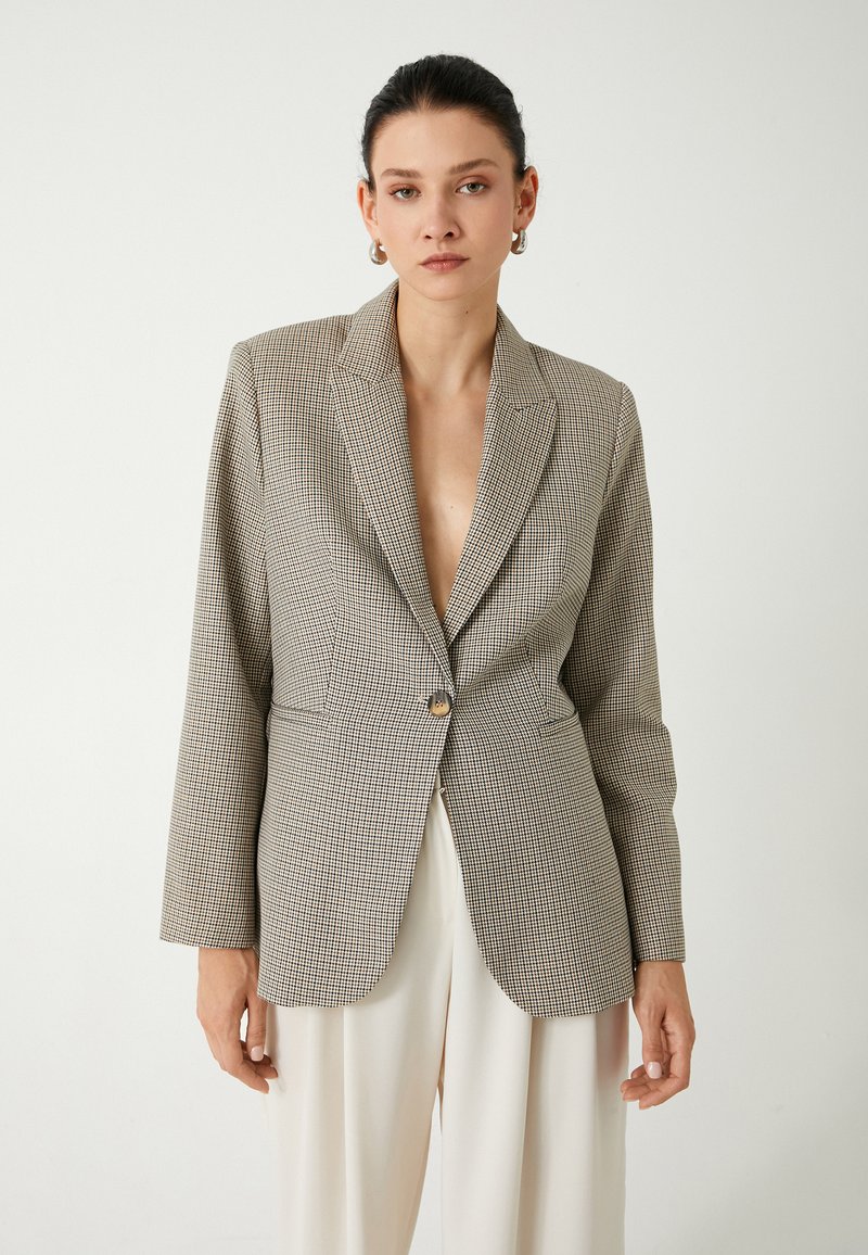 Blazer a scacchi beige e nero con chiusura a un bottone, revers a punta e maniche lunghe. Presenta una vestibilità sartoriale e una texture liscia.
