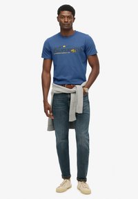 Mand iført en blå grafisk t-shirt, grå sweater bundet om livet, blå jeans, beige sneakers og et armbåndsur, står op mod hvid baggrund.