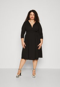 Vestido negro con escote en V pronunciado y mangas de tres cuartos, falda acampanada, hecho de tela elástica. La modelo lleva tacones de plataforma plateados.