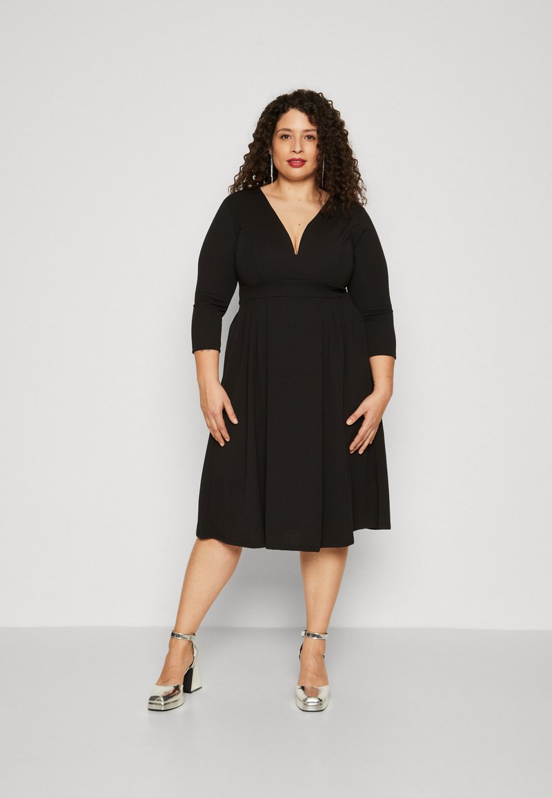 Vestido negro con escote en V pronunciado y mangas de tres cuartos, falda acampanada, hecho de tela elástica. La modelo lleva tacones de plataforma plateados.