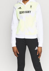 Sweat à capuche blanc zippé avec accents tie-dye vert citron, portant l'inscription « GERMANY » en noir et le logo Adidas sur la poitrine. Coupe slim.