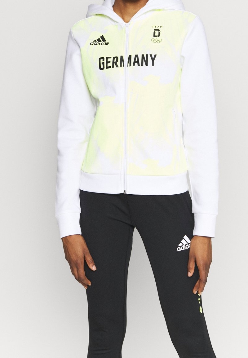 Sweat à capuche blanc zippé avec accents tie-dye vert citron, portant l'inscription « GERMANY » en noir et le logo Adidas sur la poitrine. Coupe slim.