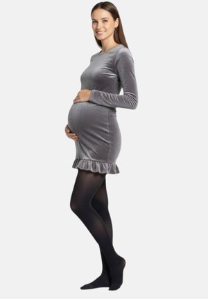 Schwangere Frau trägt ein enganliegendes graues Samtkleid und schwarze Strumpfhosen, lächelt und hält ihren Bauch vor einfarbigem Hintergrund.