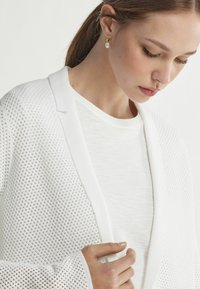 Vit cardigan med ett nätmönster och solida kanter, lagd över en enkel vit topp. Hand som försiktigt håller i kanten av cardigans.