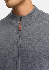 Pull gris en tricot avec un col montant, doté d'une fermeture éclair et d'une tirette en cuir. La texture est lisse et uniforme, avec des accents côtelés.
