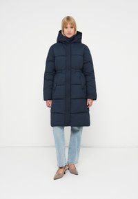Bucată de îmbrăcăminte căptușită de culoare albastru marin, cu glugă, având un sistem de închidere cu nasturi, buzunare laterale și un design până la genunchi. Purtați peste blugi de culoare albastru deschis.