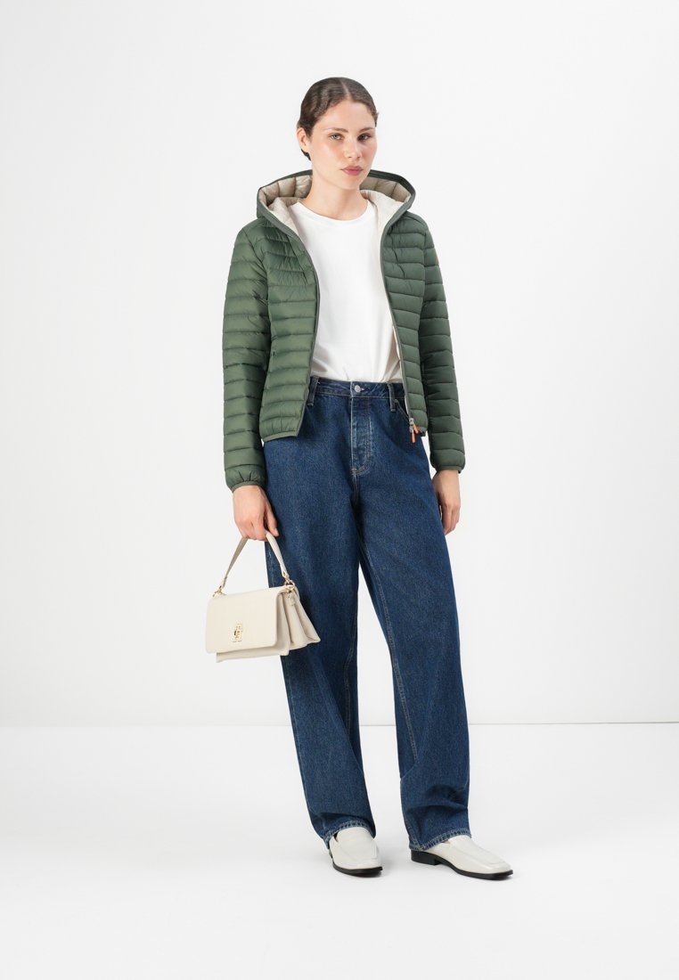 Femme portant une doudoune verte à capuche, une chemise blanche, un jean bleu ample, des chaussures blanches, tenant un sac à main beige devant un fond blanc.