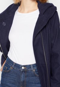 Giacca navy con zip e cappuccio, indossata sopra una maglietta bianca. Abbinata a jeans blu a vita alta. Texture liscia, design casual.