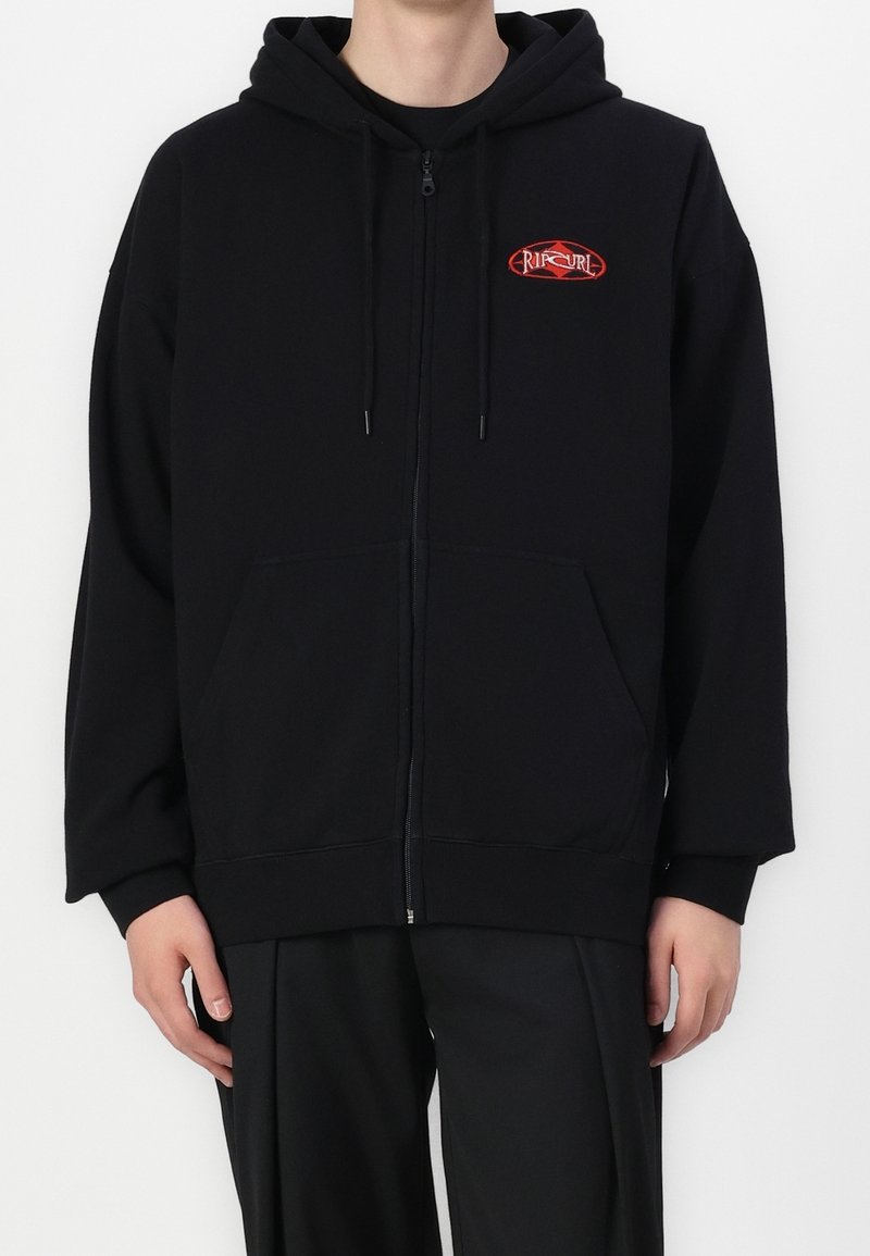 Personne portant un sweat à capuche noir zippé avec poches avant et un petit logo rouge Rip Curl sur la poitrine gauche.