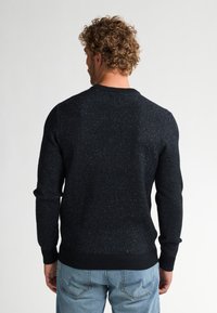 Donkerblauwe trui met een getextureerd, gevlekt patroon. Het heeft een ronde halslijn, geribbelde manchetten en een zoom, gecombineerd met lichtblauwe spijkerbroek.