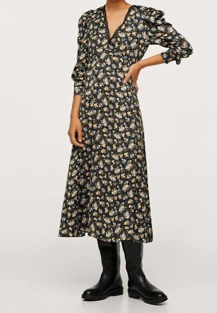 Femme portant une robe midi noire à motifs floraux avec des manches bouffantes et des bottes noires montantes jusqu'aux genoux, debout d'une main sur la hanche.