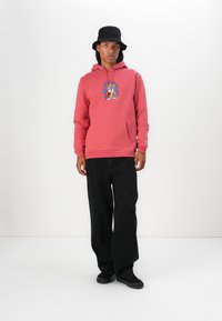Roze hoodie met een levendige grafische print op de borst, kangoeroezak en geribbelde mouwen. Gecombineerd met zwarte wijde broek en zwarte schoenen.