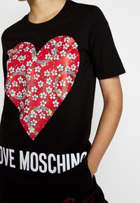 Black t-shirt featuring a large red heart graphic with floral patterns and "LOVE MOSCHINO" text. Short sleeves and crew neckline.