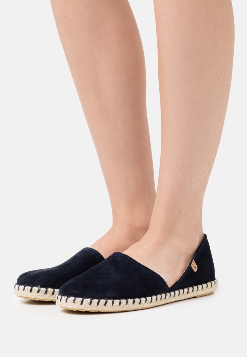Paire de jambes nues portant des espadrilles bleu marine à enfiler avec des semelles en jute tissée sur un fond clair.