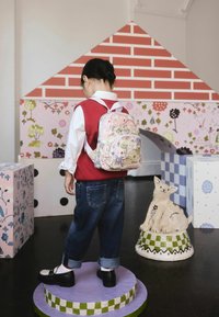 Otrok v rdeči jopici in cvetličnem nahrbtniku stoji na podstavku, v ozadju pa so barvno vzorčaste kocke in dekorativna skulptura psa.
