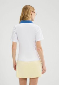 Biała koszulka polo z niebieskim kołnierzykiem, krótkimi rękawami i dopasowanym krojem. Modelka nosi żółtą spódnicę, prezentując minimalistyczny styl.
