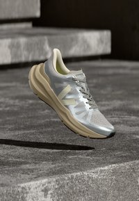 Αθλητικό παπούτσι ανοιχτού γκρι και μπεζ με κορδόνια, με επώνυμα χαρακτηριστικά, που εμφανίζεται στον αέρα πάνω από τσιμεντένια σκαλοπάτια και έδαφος.