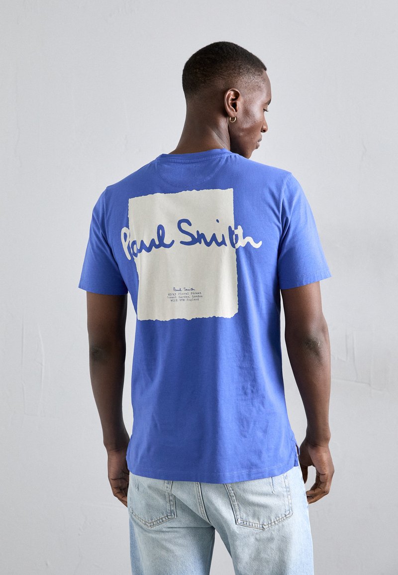 Zila kokvilnas T-krekls ar lielu baltu kvadrātu, kurā ir teksts "Paul Smith", un detaļas uz muguras. Īsas piedurknes un aplis apkaklē.