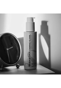 Glatte Creme-Serum in einer hohen, matten grauen Flasche mit einer weißen Pumpe, beschriftet mit "Sebastian Taming Elixir", neben einer runden Uhr platziert.