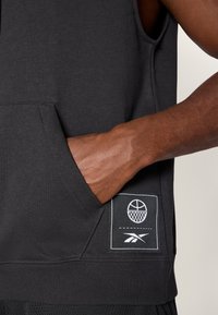 Sudadera sin mangas negra con un bolsillo frontal, tejido texturizado y una etiqueta de logo blanca con un gráfico de baloncesto y el logo de la marca.