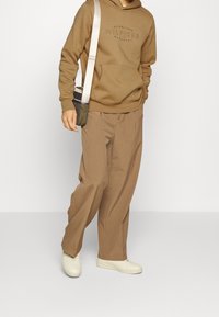 Brown hoodie with logo, loose beige trousers, and white sneakers. A small crossbody bag in dark green and tan accents is worn.