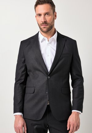 Man met kort donker haar en baard, gekleed in een zwart colbert en een wit overhemd, staand tegen een eenvoudige lichte achtergrond.