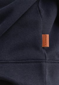 Tessuto felpa blu navy con una texture a coste, caratterizzato da una piccola etichetta rettangolare in pelle color cuoio con il marchio "GANT" impresso.