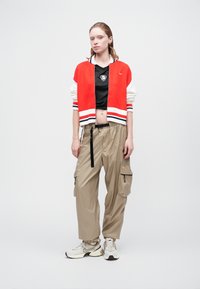 Červená a biela pletená bomber bunda, čierny sieťovaný crop top, béžové cargo nohavice a šedé tenisky. Obsahuje okrúhle logo a cargo vrecká.