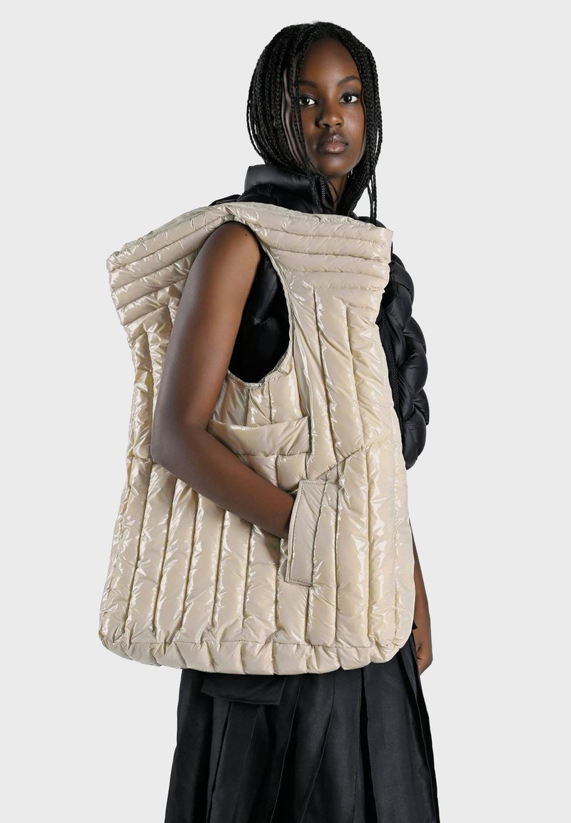 Gilet beige matelassé avec capuche surdimensionnée, poches latérales et finition brillante. Porté par-dessus un haut noir, présentant un design superposé unique.